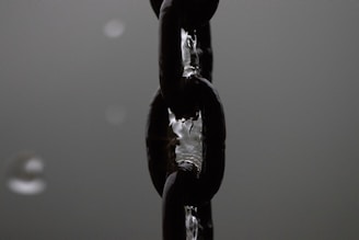 A close-up view of a sturdy metal chain, likely made of steel, with droplets of water clinging to its shiny surface. The chain appears dark and reflective against a blurred monochromatic background.