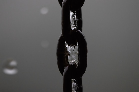 A close-up view of a sturdy metal chain, likely made of steel, with droplets of water clinging to its shiny surface. The chain appears dark and reflective against a blurred monochromatic background.