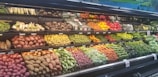 A grocery store produce section displays a variety of fresh vegetables and fruits neatly arranged on shelves. The upper shelves contain items like corn, radishes, and root vegetables, while the lower shelves are filled with potatoes, bell peppers, and carrots. A wide array of green leafy vegetables runs along the right side.