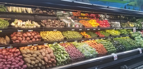A grocery store produce section displays a variety of fresh vegetables and fruits neatly arranged on shelves. The upper shelves contain items like corn, radishes, and root vegetables, while the lower shelves are filled with potatoes, bell peppers, and carrots. A wide array of green leafy vegetables runs along the right side.
