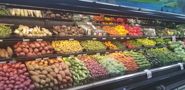 A grocery store produce section displays a variety of fresh vegetables and fruits neatly arranged on shelves. The upper shelves contain items like corn, radishes, and root vegetables, while the lower shelves are filled with potatoes, bell peppers, and carrots. A wide array of green leafy vegetables runs along the right side.