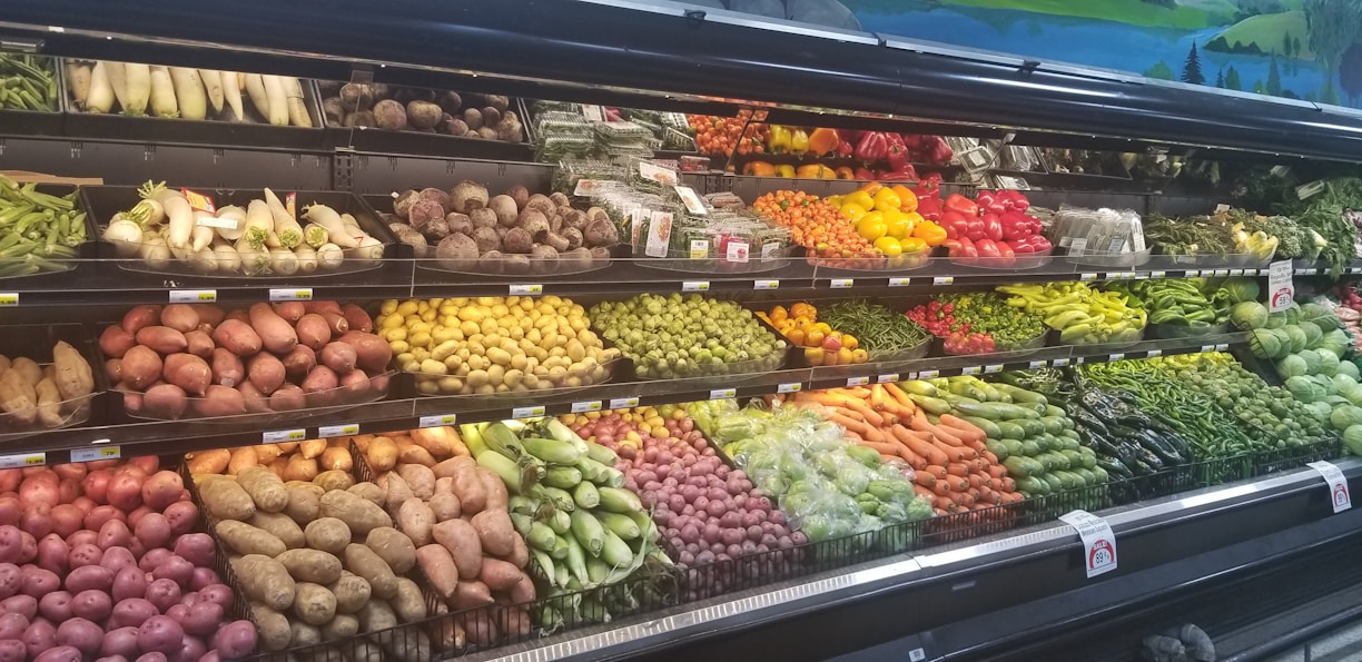 A grocery store produce section displays a variety of fresh vegetables and fruits neatly arranged on shelves. The upper shelves contain items like corn, radishes, and root vegetables, while the lower shelves are filled with potatoes, bell peppers, and carrots. A wide array of green leafy vegetables runs along the right side.