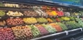 A grocery store produce section displays a variety of fresh vegetables and fruits neatly arranged on shelves. The upper shelves contain items like corn, radishes, and root vegetables, while the lower shelves are filled with potatoes, bell peppers, and carrots. A wide array of green leafy vegetables runs along the right side.
