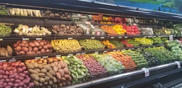 A grocery store produce section displays a variety of fresh vegetables and fruits neatly arranged on shelves. The upper shelves contain items like corn, radishes, and root vegetables, while the lower shelves are filled with potatoes, bell peppers, and carrots. A wide array of green leafy vegetables runs along the right side.