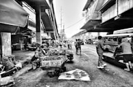 A bustling outdoor market set under an elevated train track or roadway. Vendors sit alongside their carts filled with fruits and vegetables, while people move around engaging in trade. The street is busy with pedestrians and vehicles, including the recognizable jeepney. The atmosphere is lively with a sense of daily local activity.
