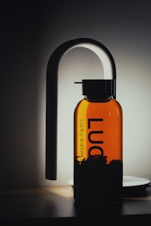 A sleek, modern glass water bottle with black lettering stands upright against a softly lit background. The bottle has a distinctive, elegant spout attachment, resembling a sophisticated dispenser or faucet, casting a gentle shadow.
