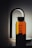 A sleek, modern glass water bottle with black lettering stands upright against a softly lit background. The bottle has a distinctive, elegant spout attachment, resembling a sophisticated dispenser or faucet, casting a gentle shadow.