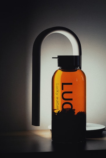 A sleek, modern glass water bottle with black lettering stands upright against a softly lit background. The bottle has a distinctive, elegant spout attachment, resembling a sophisticated dispenser or faucet, casting a gentle shadow.