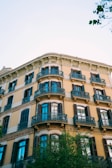 A classic European-style apartment building with multiple stories, featuring decorative iron balconies and large windows with shutters. The building's facade is adorned with intricate architectural details, and a tree extends into the frame from below, adding greenery to the scene. The sky is clear and blue, providing a bright backdrop.