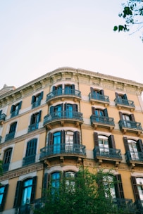 A classic European-style apartment building with multiple stories, featuring decorative iron balconies and large windows with shutters. The building's facade is adorned with intricate architectural details, and a tree extends into the frame from below, adding greenery to the scene. The sky is clear and blue, providing a bright backdrop.