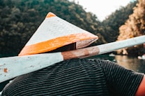 A person wearing a woven straw hat with an orange and white design is holding a wooden oar over their shoulder. The background consists of green, leafy hills and a body of water, creating a serene and natural setting. The person is wearing a striped shirt.