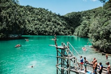 A scenic view of a turquoise lagoon surrounded by lush green forested hills. People are engaging in leisure activities such as swimming, paddling in small boats, and preparing to jump from a wooden platform. The atmosphere is lively and adventurous, with a group of people gathered near the jumping platform, capturing the vibrant outdoor spirit.