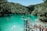 A scenic view of a turquoise lagoon surrounded by lush green forested hills. People are engaging in leisure activities such as swimming, paddling in small boats, and preparing to jump from a wooden platform. The atmosphere is lively and adventurous, with a group of people gathered near the jumping platform, capturing the vibrant outdoor spirit.