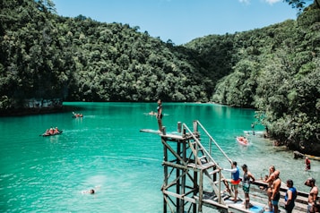 A scenic view of a turquoise lagoon surrounded by lush green forested hills. People are engaging in leisure activities such as swimming, paddling in small boats, and preparing to jump from a wooden platform. The atmosphere is lively and adventurous, with a group of people gathered near the jumping platform, capturing the vibrant outdoor spirit.