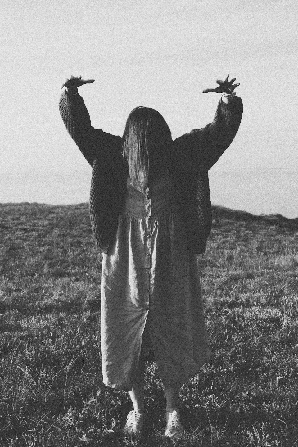 The image depicts a person standing alone on a grassy field, with their arms raised high above their head and their hands outstretched. The person is wearing a long dress that reaches down to their knees, adding an elegant touch to the scene. The background of the image shows a vast expanse of sky, providing a sense of openness and freedom.