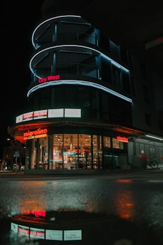 A modern building with curved architecture featuring illuminated blue LED lines against a dark night sky. The storefront, labeled 'migrolino', displays brightly lit interior with visible products through large glass windows. A reflection of the building is seen on wet pavement, enhancing the nighttime scene.