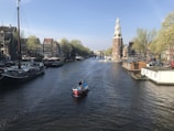 A serene canal scene with a small red boat navigating through the water. Historic brick buildings line the canal, complemented by lush green trees. A prominent tower stands on the right, overseeing the gentle flow of the canal. Several houseboats and larger moored vessels are visible along the banks, creating a picturesque cityscape.