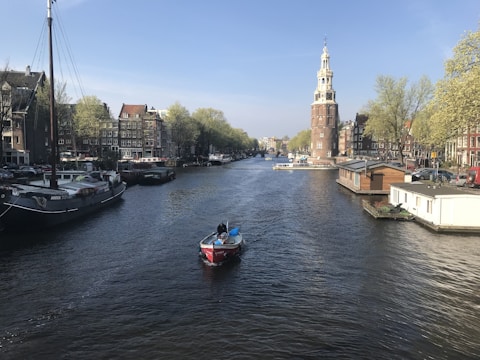 A serene canal scene with a small red boat navigating through the water. Historic brick buildings line the canal, complemented by lush green trees. A prominent tower stands on the right, overseeing the gentle flow of the canal. Several houseboats and larger moored vessels are visible along the banks, creating a picturesque cityscape.