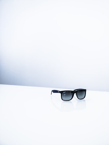 A pair of black sunglasses resting on a shiny white surface with a minimalist white background, creating a stark contrast between the dark frames and the bright surroundings.