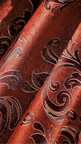 Intricate black floral patterns adorn a rich, shimmering, coppery-orange fabric. The surface has a velvety texture, with the patterns creating a sense of luxury and elegance.