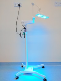 A medical standing lamp with a blue light emitting from its adjustable head, positioned inside a room with light-colored walls and a blue floor. The lamp is connected to a power outlet via a black cable, and it stands on a base with wheels for mobility.
