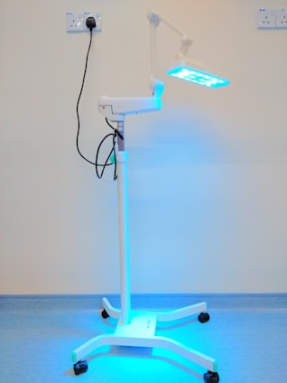 A medical standing lamp with a blue light emitting from its adjustable head, positioned inside a room with light-colored walls and a blue floor. The lamp is connected to a power outlet via a black cable, and it stands on a base with wheels for mobility.