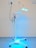 A medical standing lamp with a blue light emitting from its adjustable head, positioned inside a room with light-colored walls and a blue floor. The lamp is connected to a power outlet via a black cable, and it stands on a base with wheels for mobility.