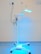 A medical standing lamp with a blue light emitting from its adjustable head, positioned inside a room with light-colored walls and a blue floor. The lamp is connected to a power outlet via a black cable, and it stands on a base with wheels for mobility.