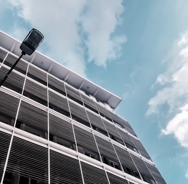 A modern building with horizontal louvers covering its windows is set against a bright blue sky with scattered clouds. A tall streetlight stands in the foreground, casting a vertical element against the predominantly horizontal lines of the building facade.