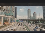 A multi-lane highway filled with vehicles is framed by tall modern buildings and ongoing construction. Overhead, an overpass runs across the image. Traffic signs and a power line tower can also be seen in the background. The sky is partly cloudy, and some greenery is visible alongside the road.