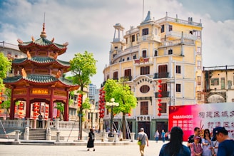 A vibrant urban scene featuring a traditional Chinese pavilion adorned with red lanterns, surrounded by a public square. An ornate, multi-story historical building with a European architectural style stands in the background. People are walking and gathering in groups, adding life to the bustling square, while a billboard with Chinese characters is visible in the foreground.
