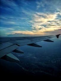 A photograph capturing the wing of an airplane during flight, set against a dramatic sky with a blend of blue and golden hues as the sun sets. The horizon is visible, with scattered clouds adding depth to the sky, and the land appears below, partially obscured by shadows.