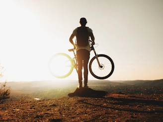Sunset silhouette of a cyclist standing beside their gravel bike on a hilltop.