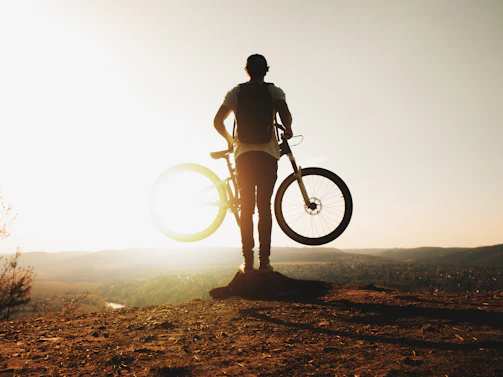 Sunset silhouette of a cyclist standing beside their gravel bike on a hilltop.
