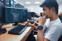 A group of people sit in front of computer monitors displaying code in an office setting. The focus is on a young person in a white shirt with a thoughtful expression, while others work in the background. The room is well-lit with white overhead lights and has a modern office design.