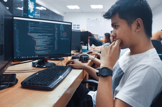 A group of people sit in front of computer monitors displaying code in an office setting. The focus is on a young person in a white shirt with a thoughtful expression, while others work in the background. The room is well-lit with white overhead lights and has a modern office design.