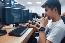 A group of people sit in front of computer monitors displaying code in an office setting. The focus is on a young person in a white shirt with a thoughtful expression, while others work in the background. The room is well-lit with white overhead lights and has a modern office design.