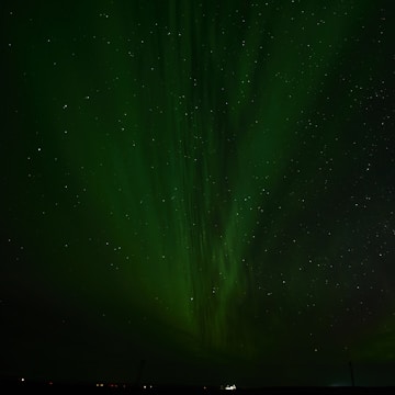 A night sky illuminated by vibrant, swirling green auroras with a scattering of stars visible throughout the scene. A faint outline of the horizon can be seen at the bottom, adding depth to the celestial display.