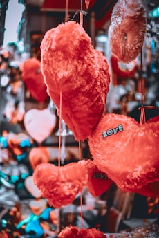 A collection of plush heart-shaped objects in various shades of red and orange hang together, some labeled with the word 'LOVE'. The background is blurred, creating a bokeh effect, with hints of other decorative items.