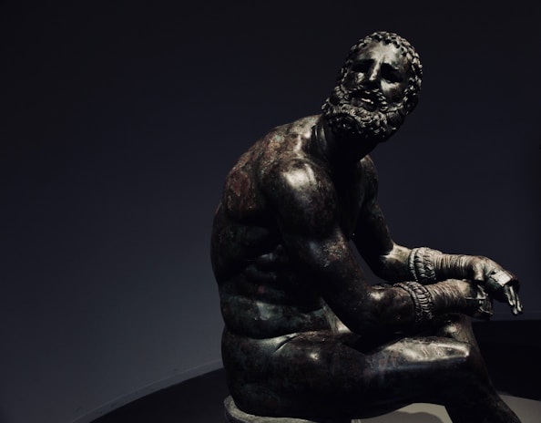 A bronze statue of a seated man with a muscular build, wearing intricate, ancient-style gloves, possibly those used for boxing. His head is turned to one side, adorned with a full beard and curly hair. The expression on his face appears contemplative and tired, in contrast to the powerful physique. His posture and the aged bronze material suggest a sense of historical significance.