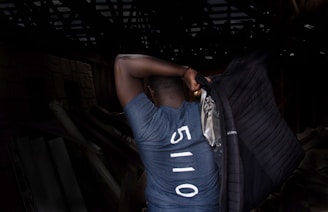 A person is seen from behind wearing a blue shirt with the number 5110 printed on it. They are holding a black jacket over their shoulder in a dimly lit environment that suggests a wooden or industrial structure.