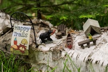 A whimsical outdoor scene with miniature objects displayed on a piece of birch bark. There is a 'Happy Trails' sign featuring colorful woodland creatures. Nearby, a small black bear figure stands on the bark. A rustic setup includes an Adirondack chair, a canoe, a tent, and a picnic table with a small green figure sitting on it. The background features lush greenery with fine details of leaves and grass.