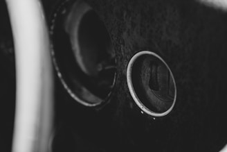 A black and white close-up image of an old steering wheel and part of a car's dashboard. The focus is on the ignition keyhole, surrounded by a circular metal ring, with textured surfaces suggesting age and wear.