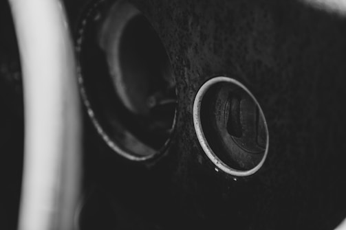 A black and white close-up image of an old steering wheel and part of a car's dashboard. The focus is on the ignition keyhole, surrounded by a circular metal ring, with textured surfaces suggesting age and wear.