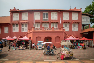 A colonial-style building painted in red with white shutters. Several stalls with colorful merchandise, including clothes and hats, are set up under pink canopies. In front of the building, a person rides a tricycle cart with an umbrella, while another person walks nearby.
