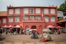 A colonial-style building painted in red with white shutters. Several stalls with colorful merchandise, including clothes and hats, are set up under pink canopies. In front of the building, a person rides a tricycle cart with an umbrella, while another person walks nearby.