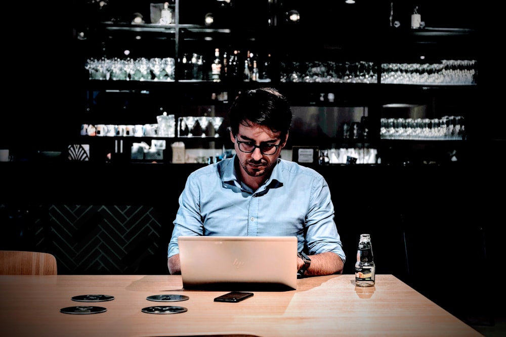 man wearing blue collared button-up long-sleeved shirt sitting and using laptop near table