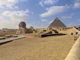 A vast desert landscape features the iconic Great Sphinx of Giza in the foreground and the Pyramid of Khafre in the background. The area is surrounded by sand and a clear blue sky, with scattered clouds adding depth to the scene. Tourists are visible along a path leading toward the structures, signifying its status as a historical and cultural landmark.