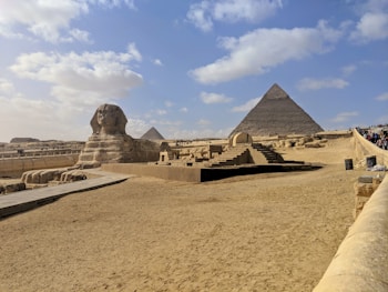 A vast desert landscape features the iconic Great Sphinx of Giza in the foreground and the Pyramid of Khafre in the background. The area is surrounded by sand and a clear blue sky, with scattered clouds adding depth to the scene. Tourists are visible along a path leading toward the structures, signifying its status as a historical and cultural landmark.