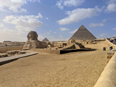 A vast desert landscape features the iconic Great Sphinx of Giza in the foreground and the Pyramid of Khafre in the background. The area is surrounded by sand and a clear blue sky, with scattered clouds adding depth to the scene. Tourists are visible along a path leading toward the structures, signifying its status as a historical and cultural landmark.